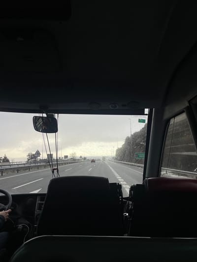 The image shows a view from the inside of a bus driving on a highway, possibly the A-6, towards Madrid. The weather is cloudy and there is light rain falling. The bus is traveling in the right lane, with other vehicles visible in the distance. The highway is divided by a white line with a shoulder on each side. The image is taken from the back of the bus, looking towards the front of the bus. The driver's seat and the steering wheel are visible to the left of the frame. The passenger seats are visible in the center of the frame. The windshield is covered in rain droplets, and the rearview mirror is positioned in the upper left of the frame. The image was taken in Guadarrama, Spain, at the coordinates 40.697, -4.127.