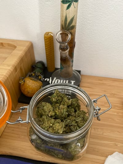 The image displays a close-up view of several objects on a wooden surface, likely indoors. In the foreground, a clear glass jar with an orange-rimmed lid is filled with dried cannabis buds. To the left of the jar, a portion of a light-colored wooden tray is visible. Behind the jar, a tall, clear glass pipe or bong decorated with green cannabis leaf imagery stands upright. Next to the bong, a textured, honeycomb-patterned yellow candle is positioned. Further to the left, near the wooden tray, is a small, yellow, fruit-like object adorned with several small, glass bee figures. In the background, a white wall is visible. The image does not provide information about the specific location within the city of Terrassa, Spain.