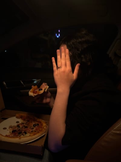 The image depicts a person in what appears to be a dark vehicle, possibly a car. The individual has dark hair and is wearing a black shirt. The person's hand is raised near their face, and a ring is visible on one of their fingers. In the foreground, there's an open pizza box with a partially eaten pizza. A single slice of pizza is being held up. The pizza appears to be topped with tomatoes and olives. The interior of the vehicle is dark, with hints of city lights visible through the window. There is no verifiable evidence of location in the city of Valchedram, Bulgaria.