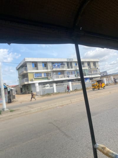 This is a street scene in Jimeta, Nigeria, captured from within a vehicle, likely a motorized rickshaw or similar open-air transport. In the foreground, a dark, textured surface, possibly the roof or canopy of the vehicle, partially obstructs the view. A dark, vertical bar, possibly part of the vehicle's structure, runs through the center of the frame.
The mid-ground features a multi-story building with a light-colored facade and blue-tinted windows. A sign on the building reads "EQUITY HOUSE." Below the building, a white fence separates it from a paved road. Several individuals are visible: a person in a red shirt walks across the road, and another person is visible near a small stall on the left side of the road. A yellow, three-wheeled vehicle, likely a "Keke Napep" or similar, is driving on the road in front of the building, carrying passengers. Another white vehicle is parked behind the fence.
The background consists of a clear sky with scattered white clouds. The overall impression is a snapshot of daily life on a street in an urban or semi-urban environment in Jimeta.