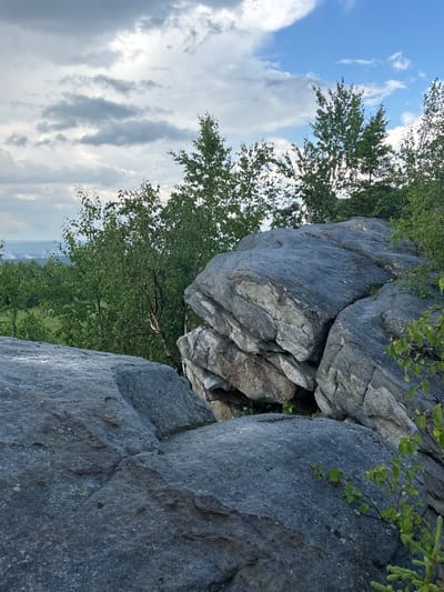 The image depicts a rocky outcrop with trees and a cloudy sky, likely taken on the Kiselovsky Ridge near the city of Gubakha, Russia. The foreground is dominated by large, grey boulders, some of which appear weathered and fractured. Lush green trees, with thin trunks and abundant leaves, grow behind and around the rocks. The sky above is a mix of grey and white clouds, with patches of blue visible. In the distance, a hazy landscape suggests rolling hills or mountains. The scene is natural and appears to be a popular spot for outdoor activities. There are no people or animals visible in the image.