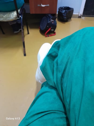 A person is seated, wearing green scrubs and white shoes, in what appears to be a room with a yellow tiled floor. In the background, a dark brown wooden desk with a metal drawer handle is visible. To the right of the desk, a black trash bag is placed on the floor, and further to the right, a red and black industrial fan is situated. A chair with a light blue cushion is partially visible on the left side of the frame. The text "Galaxy A13" is present in the lower left corner of the image, indicating the device used for capture. The location of Zaşağan, Kazakhstan is not visually discernible from the image content.