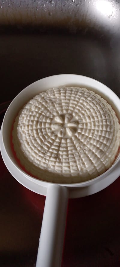In Kanelovskaya, Russia, a round, white cheese with a basket-weave pattern sits in a white cheese mold. The mold has a handle and is resting on a dark red surface. In the background, a stainless steel sink is visible with water droplets on its surface. The lighting is dim, creating a dramatic effect with highlights and shadows.