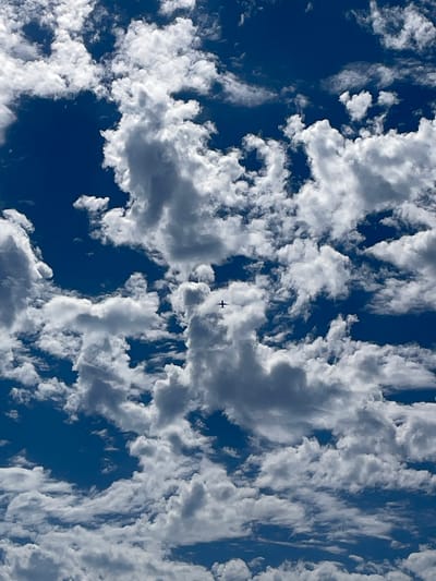 The image shows a clear blue sky filled with fluffy white clouds, some of which are shadowed, indicating the presence of sunlight. A small, dark airplane is visible flying among the clouds. The perspective is looking up from the ground. There are no people visible, nor are there any man-made structures such as buildings or roads. The image appears to have been taken during daylight hours. The location is specified as the city of Santa Monica, United States of America.