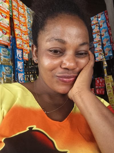 A close-up, frontal portrait captures an adult female in a retail establishment within Usiefrun, Nigeria. The subject, possessing dark skin and dark, coily hair, displays a faint, closed-mouth smile, with her left eye slightly narrowed. Her left hand is positioned under her chin, with fingers resting against her cheek and jawline. She is wearing a top that features a yellow top portion transitioning to orange, with a dark, stylized graphic at the bottom. A thin, silver-toned necklace with small beads or charms is visible around her neck, and a small, clear or silver-toned stud earring is in her left earlobe. The background comprises densely packed retail shelving and hanging displays. On the left, rows of white and orange 'PEAK CREAMER' sachets, alongside blue 'PEAK MILK' sachets, are prominently featured. On the right, blue and yellow 'GOLDEN MORN' sachets are visible, alongside red 'NESTLE MILO' packages. Numerous small bottles are arranged on shelves in the mid-background.