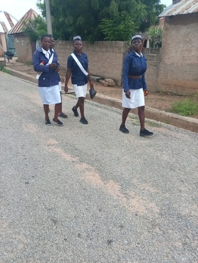 Three young women in matching blue uniforms and white skirts walk single file on a gravel road in Zinna, Nigeria. The woman in front wears a striped cap and carries a black bag in her right hand. The middle woman, also in uniform, holds a small object in her right hand. The woman farthest to the left wears a white collared shirt with a blue jacket and a white sash, and she is carrying a bottle in her right hand. A brick wall and trees are visible in the background. The sky is overcast.