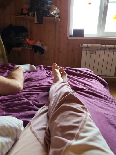 A person lies on a bed covered with a purple comforter. The person's legs are visible, one with a white sock and the other in pink pajamas. To the left of the bed, there are shelves filled with clothing. A window with a radiator beneath it is visible on the right side of the image. The walls are made of wood paneling.