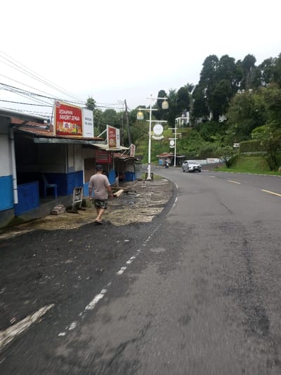 The image depicts an overcast daytime scene alongside a two-lane asphalt road, which appears damp. On the left, a series of simple, open-fronted structures with corrugated metal roofs line the roadside. Their lower walls are painted blue with white trim. Multiple commercial signs, predominantly red and white with Indonesian text ("SEDAPNYA FAVORIT SEMUA" and "WARUNG BU RUDIN") and product imagery, are affixed to these structures. Power lines are visible strung above.
A single male figure, likely middle-aged, with light skin, walks barefoot on the dark, damp dirt and gravel shoulder adjacent to the structures. He wears a light brown t-shirt and camouflage-patterned shorts, moving away from the camera towards the left. He carries a light-colored wooden object, possibly a slat or small board, in his right hand.
To the right of the road, the terrain slopes steeply upwards, covered by dense green foliage and numerous trees. A series of white, ornate streetlights with multiple decorative lantern-style fixtures and circular signs are spaced along the roadside, ascending the slope. Further up the hillside, amidst the trees, several modern white buildings are partially visible. A light-colored sedan is observed driving away from the camera on the road, with a motorcycle partially visible behind it. The presence of Indonesian language signage contradicts the specified location of Deothang, Bhutan.