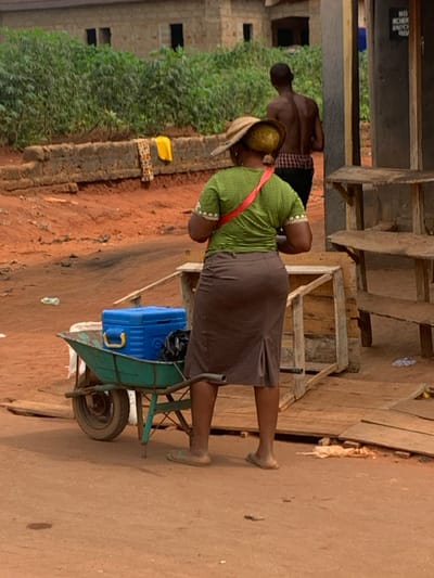 This image depicts an outdoor street scene on a reddish-brown dirt road in Aguleri, Nigeria. In the foreground, a woman stands with her back to the camera, wearing a straw hat, a green striped short-sleeved top with a red diagonal strap, a brown knee-length skirt with a back slit, and light-colored flip-flops. Adjacent to her left is a green metal wheelbarrow containing a blue cooler with a yellow handle and a black plastic bag. To her right, a multi-tiered wooden market stall constructed from light planks is visible. Further in the background, a shirtless man wearing dark pants walks away from the viewer. The mid-ground features a low concrete wall draped with a yellow fabric and a patterned fabric. Beyond the wall, dense green foliage, likely cassava plants, and an unfinished concrete building with open window frames are present. Scattered refuse is visible on the dirt ground, and a partially obscured sign on a vertical post reads "NO HOOK..."