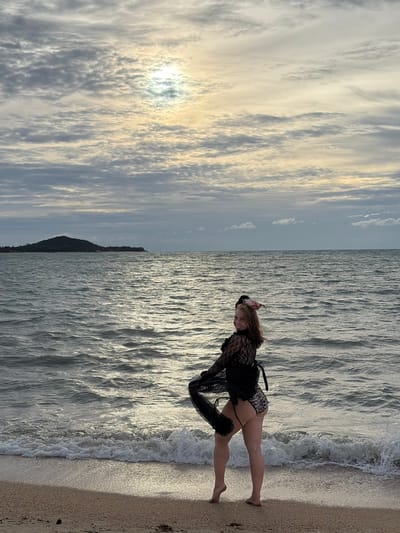 A woman stands on a sandy beach in Ko Samui District, Thailand, facing the ocean. The sky is overcast with patches of sunlight breaking through the clouds. The sun is positioned in the upper left quadrant of the frame, creating a bright halo effect. The ocean is calm with gentle waves lapping at the shore. In the distance, a dark, tree-covered island is visible. The woman is wearing a black lace top and leopard print bottoms. She is holding a black sheer fabric in her hands, which billows out behind her. Her hair is light brown and she has a pink accessory on her head. Her legs are bare and she is standing on her tiptoes. The sand on the beach is light brown and appears to be composed of small shells and pebbles.