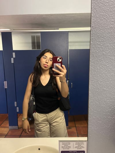 The image is a selfie taken in a bathroom, likely a public restroom, in the city of San Antonio, United States of America. A young woman with brown hair and glasses is visible, holding a smartphone with a maroon case. She is wearing a black top, khaki pants, and a gold watch. A black shoulder bag is slung across her body. The background features blue bathroom stalls, a white sink, and brown tiled flooring. A sign in English and Spanish with a domestic abuse hotline number is visible on the right side of the frame. An air vent is visible in the background.