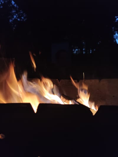 A fire burns brightly in a dark, outdoor setting, possibly a backyard or park in Kolpakovo, Russia. The flames, rendered in shades of orange, yellow, and hints of blue, rise from a dark metal fire pit. The background is obscured by darkness, with faint silhouettes of trees and what appears to be a blurry, indistinct figure or object in the upper right quadrant, possibly a person or a reflection. The overall impression is of a nighttime scene, with the fire being the sole, vibrant source of light.