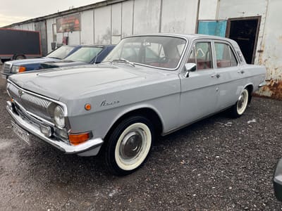 The image shows a grey GAZ-24 Volga parked on a wet gravel surface in what appears to be a parking area or open lot. The car is a four-door sedan, exhibiting a chrome grille, white-walled tires, and a partially visible license plate. To the left of the Volga is a dark-colored Mercedes-Benz sedan, and further to the left is a black trailer or truck bed.

Behind the vehicles is a grey building with vertical panels, possibly a warehouse or garage. A sign with Russian text and a phone number is visible on the building. An open doorway or garage opening is present on the right side of the building. The sky is overcast, indicating a cloudy day.

Based on license plate and text, location of the image is Khar'ino, Russia.
