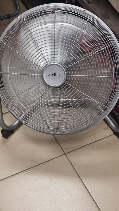 A close-up, slightly overhead shot shows a silver industrial fan with a circular grill and a central hub bearing the brand name "aceline" in black lettering. The fan is positioned on a tiled floor, which appears to be made of large, light-colored square tiles with dark grout lines. The fan's stand, made of gray metal, is visible in the lower right and lower left corners of the image. In the background, behind the fan, a dark object and some shiny metal tubing can be seen, suggesting an indoor industrial or workshop setting. The lighting is even, without strong shadows, hinting at artificial light. No people are present in the image. The overall impression is of an object at rest in a functional environment.