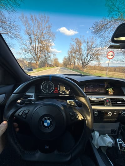 The image is a first-person perspective from inside a BMW car, presumably on a road trip in Gorna Rositsa, Bulgaria. The steering wheel, with the BMW logo and "M" badge, dominates the foreground. The dashboard is visible, featuring a screen displaying radio stations and car information (such as time, temperature, and odometer reading). A small Yoda figurine sits on the dashboard. Through the windshield, a road stretches into the distance, flanked by bare trees against a blue sky with some clouds. A road sign is also visible. A partially visible hand is gripping the steering wheel. A white cloth rests on the center console.