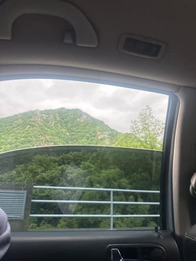 The image shows a view from inside a vehicle, likely a car, looking out through a window. The window is partially open, revealing a landscape featuring a mountain covered in green vegetation under an overcast sky. A metal fence or barrier is visible close to the window, partially obstructing the view. Parts of the vehicle's interior are in the foreground, including the ceiling, a grab handle, and the side panel. The location is likely near Krichim, Bulgaria, as specified in the prompt.