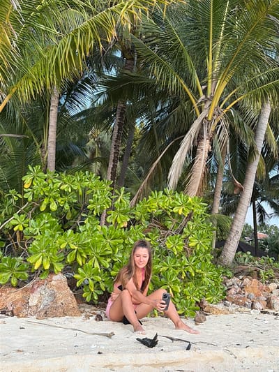 A woman is sitting on a sandy beach in Baan Taling Ngam, Thailand, with lush green foliage and palm trees behind her. She is wearing a pink bikini top and pink shorts. The woman is holding a smartphone in her right hand and looking at it. The sand is light-colored and appears to be a beach, with some rocks and driftwood scattered around. The foliage behind her is dense and vibrant green, with several palm trees towering above. The overall scene suggests a tropical beach setting.