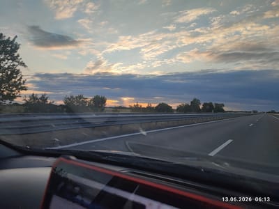 The image shows a view from inside a vehicle traveling on a highway at sunrise. The sky is filled with clouds, some of which are illuminated by the sun, creating a colorful and dramatic display. The highway stretches into the distance, with guardrails lining the sides. In the foreground, the dashboard and windshield of the vehicle are visible, with a reflection of the sky on the windshield. The location is identified as Voisil, Bulgaria, with the timestamp "13.06.2026 06:13" visible in the lower right corner. The scene is calm and serene, conveying a sense of motion and the start of a new day.