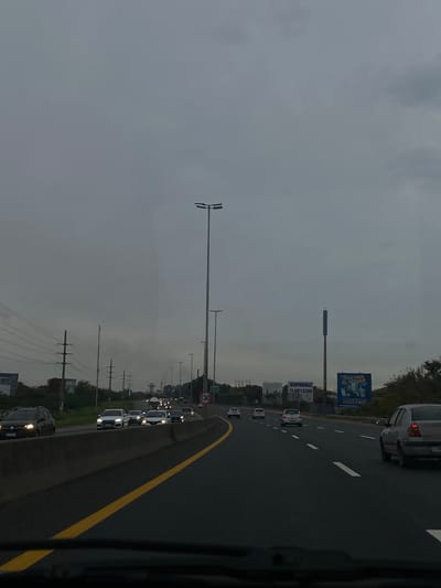 The image shows a multi-lane highway in Bernal Este, Argentina, likely taken from inside a vehicle. Cars are visible in both directions with light traffic. Overhead street lights and power lines line the side of the road. Billboards stand in the distance. The sky is overcast. A speed limit sign indicating "130" is visible.