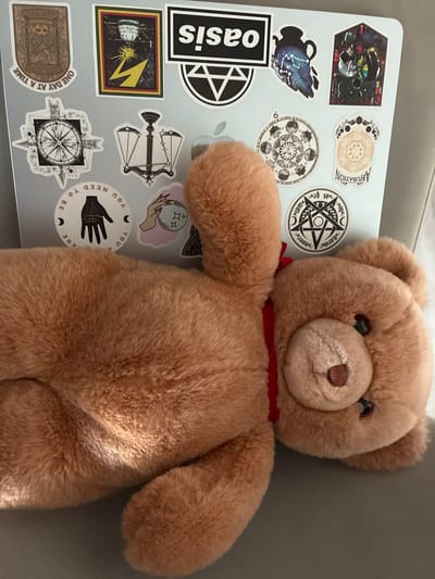 The image shows a close-up, overhead view of a light brown teddy bear placed on top of a laptop. The laptop is covered in various stickers, primarily with occult, astrological, and symbolic imagery. Some of the visible stickers include a stylized compass rose, a set of scales, a hand with an eye, astrological charts, a silhouette of a pitcher with constellations, and a black and white "Oasis" logo. A red ribbon is tied around the teddy bear's neck. The lighting suggests it's daytime, with sunlight casting shadows on the teddy bear's fur. The background is a plain, dark gray surface, possibly a couch or chair. The overall impression is one of a personal item, perhaps a workspace or a personal area, with a whimsical and slightly mystical aesthetic due to the sticker choices. The OCR text visible on the stickers includes "ABJURATION," "harme," "ah sp," "YOU NEED TO BE," and "ONE DAY AT A TIME."