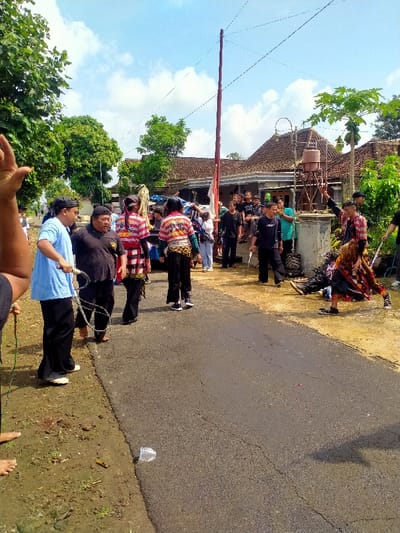 The image depicts a scene in Kecamatan Kedawung, Indonesia, located at latitude: -7.52, longitude: 111.028. A group of people are gathered on a paved road in front of a traditional Indonesian house. The road is dusty and there is a small water tank to the right of the house. Several of the people are dressed in traditional costumes, including red and white striped shirts and black pants. One person holds a whip. The person in the foreground is wearing a blue shirt and black pants. There is a plastic cup lying on the ground. The people are standing in front of the house, possibly engaged in a ceremony or traditional dance. The sky is blue with some white clouds. The houses are made of brick and concrete and the buildings are surrounded by trees.