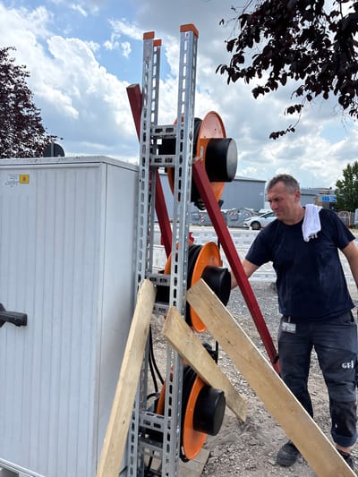 This is a medium shot, outdoor photograph of a man working on some equipment. The man, who appears to be middle-aged with short, graying hair, is wearing a dark blue t-shirt and dark pants. He has a white towel draped around his neck. He is leaning over some orange cable spools that are mounted on a silver metal rack. A red wooden plank is positioned diagonally across the spools, and another wooden plank is resting on top of the rack. The background shows a gravelly surface with some buildings and trees visible. The sky is cloudy with patches of blue. The lighting suggests it is daytime. There is some text visible on the man's pants which appears to be a logo or insignia. There is also a yellow sticker with text on the white container in the lower right of the frame. The location is identified as Mannheim, Germany. The overall impression is that of an industrial or construction site.