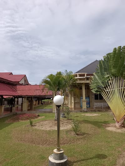 A paved walkway with red tiled roofs and beige supporting pillars is visible to the left of the image, with a person walking away from the camera down the walkway. In the foreground, a lamp post with a spherical light fixture stands in a grassy area. Behind the walkway, a building with a dark grey roof and beige walls is visible. A large traveler's palm tree with fan-shaped leaves is situated to the right of the building. The sky above is cloudy. The location is the city of Mesjid Reuntoh, Indonesia.
