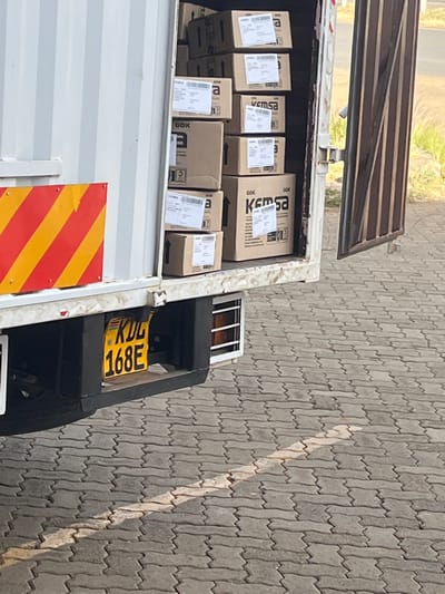 A partially loaded delivery truck has its back hatch open. Brown cardboard boxes, some labeled "KEMSA," fill the cargo space. Each box has a white label. The truck's yellow license plate reads "KDL 168E". A red and yellow striped warning marker is affixed to the truck's exterior. The scene takes place outdoors on a gray tiled surface.