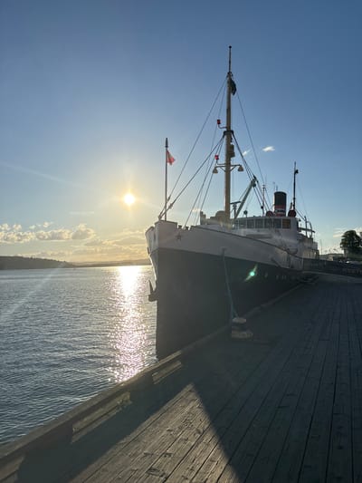 The image shows a sunlit scene at a dock in Tønsberg, Norway. A large, dark-hulled ship with white upper decks is docked to the right of the frame. The ship has several masts and rigging, with flags visible. The sun is low in the sky, casting a bright glare on the water and creating long shadows on the wooden planks of the dock. The water is calm with gentle ripples reflecting the sunlight. In the background, a hilly, tree-lined coastline is visible under a clear blue sky with a few scattered clouds. The dock itself is made of weathered wooden planks. No people are visible in the image. The scene appears to be a still, late afternoon or early evening moment.