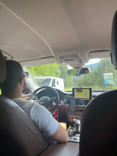 The image shows the interior of a car, looking forward from the back seat. A man is driving, wearing a white t-shirt and sunglasses. He has a beard and is looking ahead. The car's dashboard is visible, with a navigation screen displaying a map and directions. Outside the car, there are trees and a bright sky, suggesting a daytime outdoor setting. A white van is visible on the road in front of the car. The car's interior features leather seats and the Audi logo on the steering wheel. A small wooden pendant is hanging from the rearview mirror. There are also some stickers on the windshield. Based on the context of the surroundings, the car appears to be driving in or around Oberammergau, Germany, a town known for its scenic Bavarian landscapes and forests.