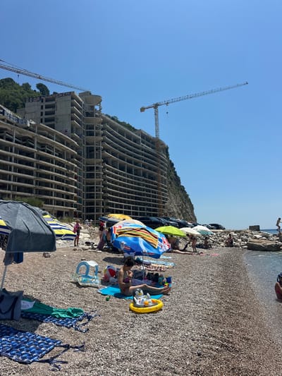 The image depicts a busy beach scene in Mirište, Montenegro, with an unfinished concrete building and construction cranes dominating the background. The building, a multi-story structure with exposed floors and pillars, is partially covered in graffiti. Several large construction cranes are visible, indicating ongoing work or abandonment.
In the foreground, beachgoers are scattered across the pebble beach, seeking shade under colorful umbrellas. Several people are sunbathing, while others are engaged in casual conversation. Children's toys, including inflatable floats and a beach ball, are present on the ground, suggesting family activities. Beach towels and mats are spread out for relaxation.
The sea is visible to the right, with some individuals in the water. The overall environment is a mix of construction and leisure, with the imposing unfinished building juxtaposed against the serene beach setting. The sky is clear and blue, typical of a sunny day.
