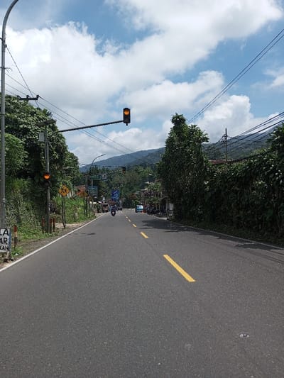 This image depicts a street scene in Boommlog, Bhutan, under a partially cloudy sky. The primary subject is an asphalt two-lane road receding into the distance. The road features yellow dashed lines marking the center and a solid white line on the left shoulder. On the left side of the road, a gray utility pole supports multiple overhead wires and a street light with an attached solar panel. A traffic light mounted on an overhead arm displays an amber light, with a second amber traffic light visible further down the road. Green vegetation, including trees and dense bushes, lines both sides of the road. A yellow diamond-shaped sign with black symbols, likely a warning sign, is visible on the left. Further down the road on the left, a blue banner or sign is partially visible amidst buildings. On the right side, lush green foliage and additional utility poles with numerous overhead wires are present. Shadows are cast across the right side of the road. Multiple parked motorcycles/scooters and a light blue van are visible further down the road on the right, near several structures. A single person on a dark-colored scooter travels away from the camera in the left lane. In the background, green, forested mountains are visible beneath the cloudy sky. A reflective road stud is present in the lower right foreground of the pavement.