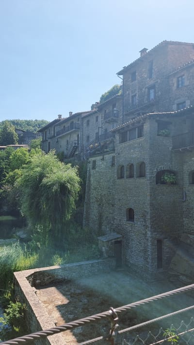 This is a vertical, eye-level, exterior shot of a stone building in Rupit i Pruit, Spain. The building is constructed of rough-hewn grey stone blocks with a terracotta tile roof. It has multiple levels with balconies, arched windows, and small balconies with wrought-iron railings. Green foliage, including trees and ivy, grows around and on the building. In the foreground, a stone wall encloses a courtyard area that is partially in shadow. A cable railing runs across the bottom of the image. The sky is clear and blue.