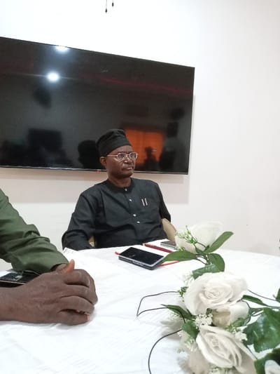 The image shows a scene inside a building in Yola, Nigeria, located at the coordinates 9.198 latitude and 12.49 longitude. The scene features two men seated at a white table covered with a white tablecloth. The man on the right, wearing a black long-sleeved shirt, black cap, and eyeglasses, is sitting upright and looking directly at the camera. The man on the left, wearing a green long-sleeved shirt, is looking down at his hands. The man on the left's hands are placed on the table, with fingers curled. There is a black cell phone, a red folder, and a black notebook on the table. A bouquet of white roses with green leaves is positioned on the table in front of the man on the right. A large black TV screen is mounted on the white wall behind them. The scene is well-lit and appears to be a formal meeting or interview setting.