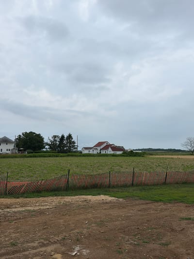The image, taken in Mattituck, USA, depicts a landscape under a cloudy sky. The scene is divided by a fence made of thin, vertical posts with orange netting. To the left of the fence is a field of green grass. Beyond the grassy field, several houses with red roofs are visible, interspersed with trees. One house, a white structure with a prominent red roof, stands out. To the right of the fence, the ground is a mix of dirt and sand, suggesting a construction or undeveloped area. In the distance, a treeline and hints of water can be seen, along with a sliver of sky at the very top. The overall impression is a rural or semi-rural setting, possibly on the outskirts of a town, under overcast conditions. No people or specific activities are evident.