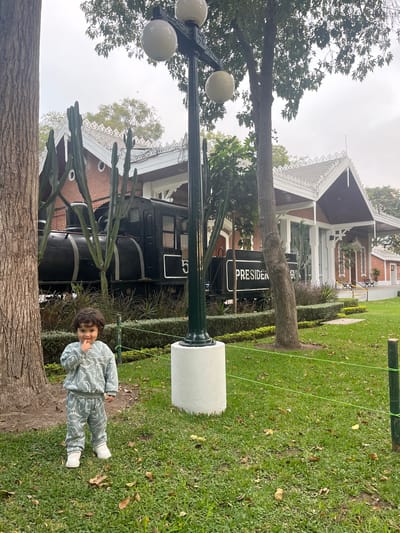 A small child is standing on grass, near a tree and a pole supporting a cylindrical, white object, possibly a speaker. The child is wearing a gray dinosaur-patterned outfit and white shoes. Behind the child and to the left is an old black train car with the word "President" partially visible on the side. A red brick building with white trim and a gabled roof is behind the train car. The photograph appears to be taken at an angle, giving the impression of a tilted scene. Based on the visual features of the building and the old train, this image might be taken in Miraflores, Peru.