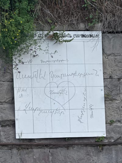 A white sign with a grid of handwritten text is mounted on a stone wall. The sign appears to be outdoors, with green vegetation and dry brush growing around it. The text on the sign is in Armenian. At the top, there is a heading that reads "ՀԱՅԱՍՏԱՆԻ ԸՆՏՐՈՒԹՅՈՒՆՆԵՐԻՆ" which translates to "To the Elections of Armenia". Below this, there are several lines of handwritten names and messages. In the center of the sign, a heart shape is drawn. The sign is attached to the wall with visible screws and washers. The stone wall is made of large, rough-hewn blocks. This sign is located in the city of Dilijan, Armenia, likely a public space or monument.