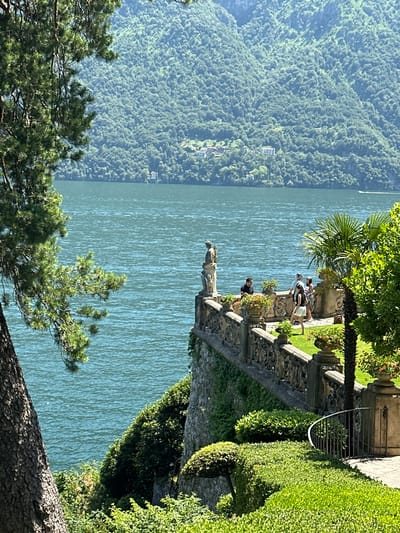 The image depicts a scenic view of Lake Como from the gardens of Villa del Balbianello in Tremezzina, Italy. In the foreground, a large pine tree on the left frames the scene, with its branches extending across the upper left quadrant. A stone balustrade with intricate carvings runs along the edge of a terrace overlooking the lake. Several potted plants with red and orange flowers are placed on the terrace, adding color to the scene.
On the terrace, a group of tourists is visible. Two men and two women are standing and conversing near the balustrade. One woman, wearing a white skirt and a black top, is walking away from the camera. A stone statue of a figure stands near the edge of the terrace, looking out at the lake. In the background, the deep blue waters of Lake Como stretch out to meet the lush green mountains that rise steeply from its shores. Several houses are scattered across the mountainside, some nestled amongst the trees. A small boat is visible on the lake in the distance, leaving a faint wake. The overall impression is one of serene beauty and classical elegance.