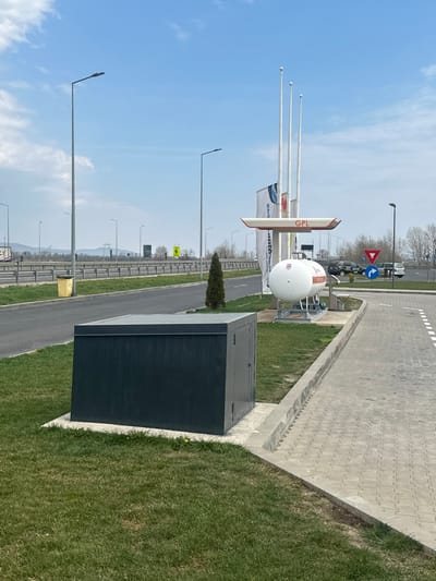 The image depicts an outdoor scene at an autogas fueling station adjacent to a highway, located in Orăștie, Romania. The sky is light blue with scattered white clouds, indicating clear weather conditions during daylight hours.
In the foreground, a patch of green grass occupies the left and bottom sections. Situated on this grass is a dark grey, rectangular utility or storage box, mounted on a concrete slab. To its right, a paved area constructed from interlocking light grey pavers features white painted lines, delineating a path or parking space. A small, conical evergreen tree stands on the grass between the utility box and the fueling station.
The midground features the autogas fueling station. A prominent structure consists of a red and white horizontal canopy with "GPL" (LPG) written in red, supported by three tall, white poles, which resemble flagpoles. Beneath this structure, a large white cylindrical liquefied petroleum gas (LPG) storage tank, labeled "GPL" and "autogas" in orange, rests on a metal platform. A black fuel dispenser is partially visible to the right of the tank.
To the left of the fueling station, a two-lane paved road runs horizontally, bordered by a metal crash barrier. Numerous tall, grey streetlights are positioned along this road and extending into the background. A yellow garbage bin is visible near the left edge of the road. Vehicles, including passenger cars and a white van, are visible on the road and in a distant parking area, but no individuals are identifiable. A red triangular "yield" sign and a blue circular "mandatory direction" sign are present near the fueling station entrance. A yellow rectangular sign and a distant sign with a Ferrari logo are visible across the highway.
The background shows a distant landscape with a line of bare trees, possibly indicating early spring or late autumn, and hills or mountains beneath the horizon. Power lines or infrastructure are also faintly visible in the far distance.