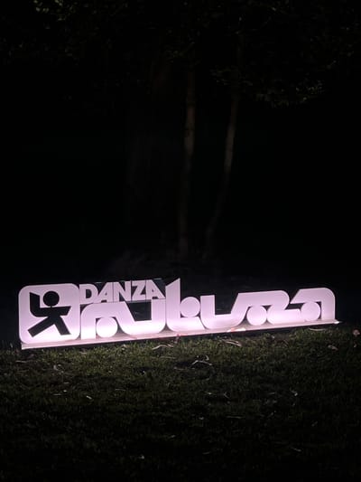 A backlit sign with the word "DANZA" and a stylized figure is positioned outdoors on a grassy area at night. The sign is white with a pinkish hue, and the letters are three-dimensional. The figure on the left appears to be a stick-like person in a dynamic pose. The sign is resting on a flat surface and is illuminated from within, casting a glow on the surrounding grass. In the background, out-of-focus trees are visible against a dark sky. The image appears to be taken in Viana do Castelo, Portugal, though no specific landmarks are identifiable.