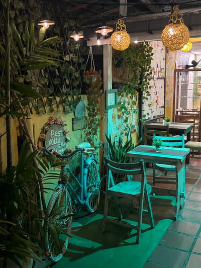 The image depicts an outdoor seating area of a restaurant or cafe, possibly located in Galle, Sri Lanka, as suggested by the decorative signage. The scene is illuminated by warm, ambient lighting from decorative lamps hanging overhead, casting a greenish hue across the space. In the foreground, a vintage blue bicycle is parked against a yellow wall adorned with vines and a circular sign featuring floral decorations and the text "Best Kottu in the Island." Several wooden tables and chairs are arranged in the seating area, with one table in the mid-ground set with a small potted plant. The floor is tiled. The overall atmosphere is cozy and inviting, with a touch of rustic charm.