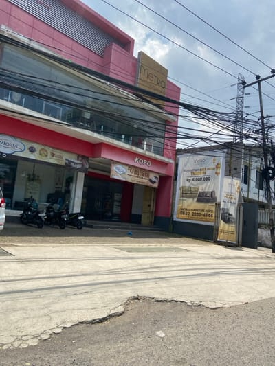 The image shows the front of a furniture store called "Metro Furniture Kopo" located in Kecamatan Margahayu, Indonesia (latitude: -6.962, longitude: 107.578). The store has a large, red-colored storefront with a glass front and a sign above the entrance that says "Metro Furniture Kopo". The store's sign also states the phone number "0882-2033-4844". There are three motorcycles parked in front of the store. There are several large signs, some on the storefront, and some on the ground. One sign advertises a "Ramadhan Sale", with a sofa pictured on the sign, for the price of "Rp. 6.000.000". The sign also shows a "trade in" option. There are multiple electrical wires strung over the storefront, and a power line tower in the background. The street in front of the store is paved and appears to be in need of repair. The environment is daytime and appears sunny.