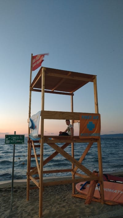 A wooden lifeguard tower stands on a sandy beach in Perea, Greece, near the Thermaic Gulf. A young boy sits cross-legged on the platform of the tower, looking towards the camera. A red flag with white lettering is affixed to the top of the tower. A sign in Greek, reading "ΣΗΜΕΙΟ ΔΕΙΓΜΑΤΟΛΗΨΙΑΣ" (Sampling Point), is visible to the left of the tower. An orange banner with a blue cross and the words "FIRST AID" and "ΠΡΩΤΕΣ ΒΟΗΘΕΙΕΣ" (First Aid) is displayed on the right side of the tower. Two paddleboards are visible on the sand near the base of the tower. The sea is calm with gentle waves, and the sky is clear with a hint of sunset colors.