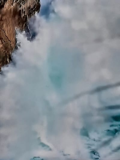 The image depicts a close-up view of a turbulent body of water, likely a river or sea, with a rocky, textured cliff face on the left. The water exhibits a range of colors from white foam and spray to pale blue and darker grey-blue hues, indicating movement and depth. The cliff is predominantly brown and grey, with visible cracks and rugged formations. Wisps of white mist or spray are obscuring parts of the cliff and blending with the water's surface. The overall impression is one of natural forces at play, with the water actively interacting with the landmass. The location is identified as Sambir, Ukraine.