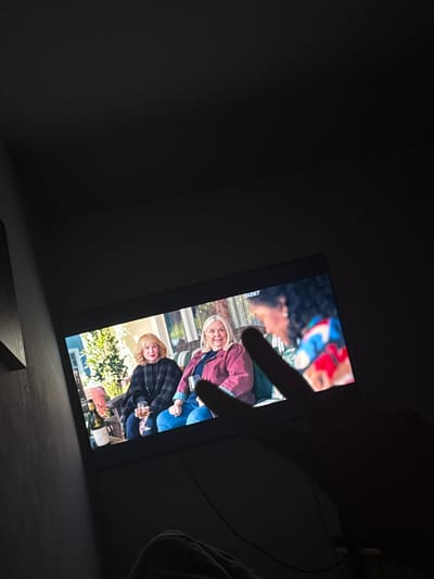 A television screen displays a scene of three women sitting on a porch. The woman on the left has blonde hair and is wearing a black and grey plaid jacket. The woman in the middle has blonde hair and is wearing a maroon jacket over a blue shirt. The woman on the right has dark curly hair and is wearing a colorful jacket with red, blue, and white patterns. One woman is holding a glass. A finger is obscuring part of the screen in the foreground. The television is in a dark room, with part of a wall visible on the left side of the image. There are no elements in the image that suggest a specific location in Neubrandenburg, Germany.