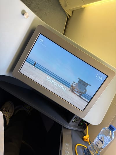The image shows a close-up view of an airplane's in-flight entertainment system screen, displaying a "Welcome" message and a beach scene with a lifeguard tower. Text on the screen indicates "8K World Business Class," "LAX Los Angeles," and "Ocean Front Walk, Pacific Palisades." A bottle of SPA Reine mineral water is visible in the foreground, next to a charging cable. In the lower left corner, a person's leg and foot, wearing a sneaker, are visible. The setting appears to be inside an airplane cabin, with the bright lighting and seating arrangement characteristic of premium class travel. There is no indication of the location being Badhoevedorp, Netherlands.