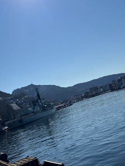 The image shows a view in Bergen, Norway. The scene includes a body of water with ripples, likely the harbor, with a mountainous backdrop lined with buildings and structures. A naval vessel, potentially docked, is visible, along with parts of city infrastructure. The sky is clear and blue. The general atmosphere suggests a sunny day. The angle of the shot indicates it was taken at a tilt.