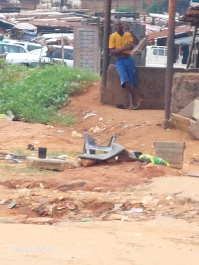 In Awkuzu, Nigeria, a young man in a yellow t-shirt and blue shorts stands leaning against a brown, textured wall. The environment appears to be an informal settlement or a semi-developed area, characterized by rough terrain and scattered debris. Behind him, a white van is visible, along with other structures suggesting an open-air market or residential area with makeshift constructions. The ground is predominantly red earth, with patches of green vegetation in the background. Scattered on the ground are various objects, including what appears to be a discarded plastic chair, and some plastic bags. The overall impression is one of a utilitarian and somewhat untidy urban setting.
