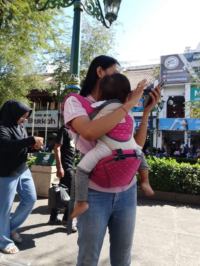 This image captures a bright, sunny day in Yogyakarta, Indonesia, likely in a busy urban area given the presence of shops and a street lamp. The primary subjects are people walking outdoors. In the foreground, a woman is carrying a baby in a pink and grey front carrier. She is looking at her phone, suggesting she might be navigating or communicating. The baby's small foot is visible, peeking out of the carrier.
To the left, another woman wearing a black hijab and glasses is also looking down, possibly at her phone or something in her hands. A man in a black t-shirt walks behind them, carrying a large black plastic bag. In the background, several other individuals are visible, some sitting down.
The scene is set along a paved walkway with some greenery and shops lining the street. Visible text includes "TOKO MAS Berkah" (Gold Shop Berkah), implying a commercial area. Other text on signs in the background relates to "KOPI POKOKE BLANGKON" (a coffee shop) and "GO SWEAT GO ION" (likely an advertisement for a drink or sports-related product). The overall atmosphere appears to be one of everyday life and commerce in a bustling Indonesian city.