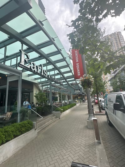 The image shows a street scene in Burnaby, Canada, specifically in front of Earls Kitchen + Bar at Station Square. A sidewalk made of light grey interlocking bricks runs along the right side of the image. A white van is parked on the street to the right of the sidewalk. Across from the van and sidewalk is a restaurant with a glass facade and a green tinted glass awning supported by dark grey metal beams. The restaurant's sign, "Earls KITCHEN + BAR", is visible. A red banner hangs from a pole, reading "STATION SQUARE". Trees with green foliage line the sidewalk, and hanging flower baskets adorn a pole. People are visible on the sidewalk and near the entrance of the restaurant. In the background, buildings are visible. The sky is overcast.